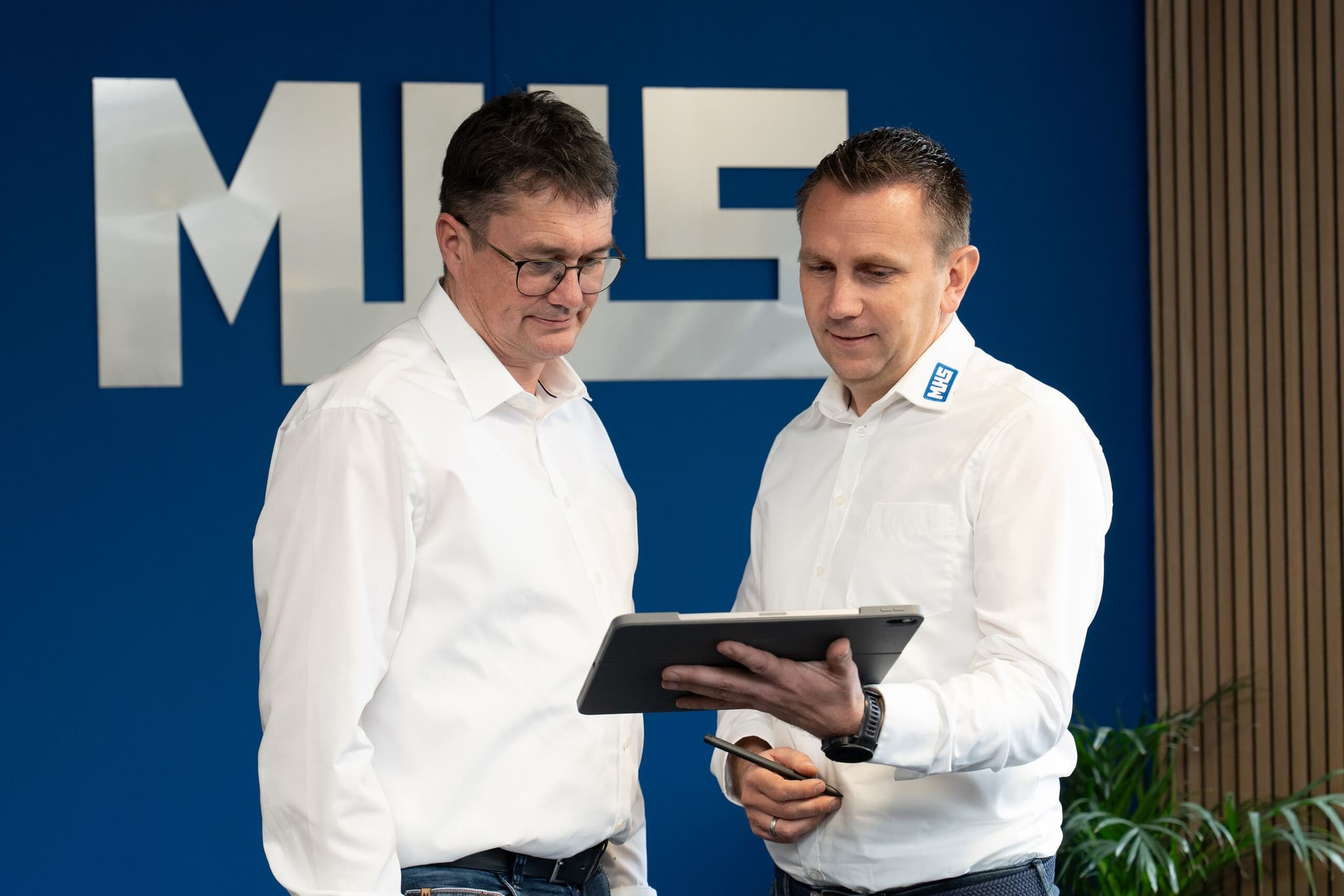 Zwei Mitarbeitende der MHS GmbH besprechen einen Auftrag am Tablet vor der Firmenwand